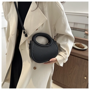 Detail of black pleated crossbody bag with gold hardware worn over a beige trench coat