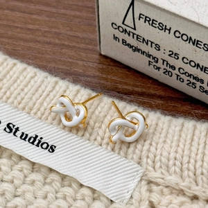 Close-up of golden knot earrings with white enamel finish on a soft knitted fabric