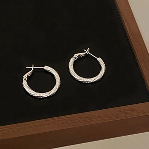 Close-up of small silver textured hoop earrings with a metallic woven pattern finish