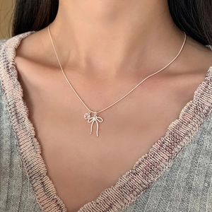 Silver bow necklace with pink heart pendant worn by a model on a grey sweater background