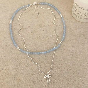 Collier double rang avec perles bleues et pendentif nœud argenté sur fond en lin naturel