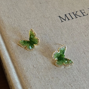Detailed close-up of green crystal butterfly stud earrings with gold plating on linen