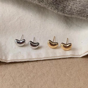 Minimalist bean-shaped stud earrings in polished gold and silver on soft fabric background