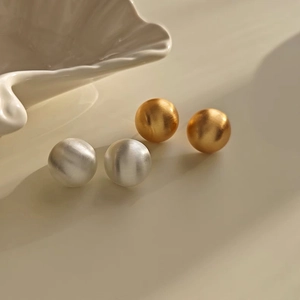 Minimalist matte gold and silver round stud earrings shown on a premium cream stand