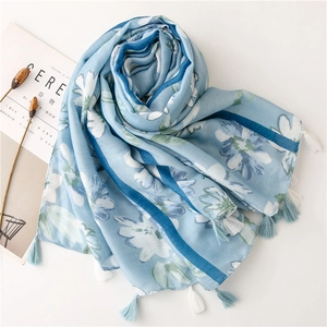 Dettaglio della sciarpa azzurra con stampe floreali bianche e nappe su sfondo bianco pulito