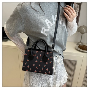 Detail of a black mini square bag featuring a vibrant red cherry print and top handle