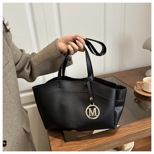 Detail of matte black leather tote bag featuring a gold M-pendant and dual handles