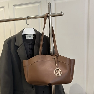 Large brown leather tote bag with metal M pendant hanging on a garment rack