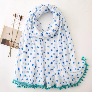 White polka dot scarf with blue pom-pom fringe trim shown in a flat lay minimalist setup