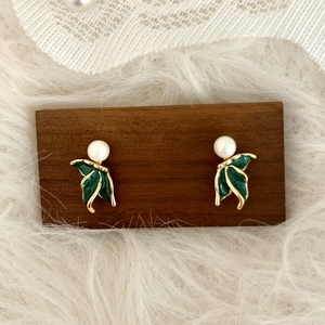Elegant fishtail pearl stud earrings with green enamel leaves on a wooden display stand