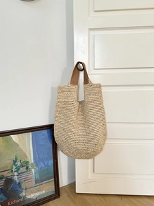 Detail of woven straw tote bag with brown leather handles hanging on a white door