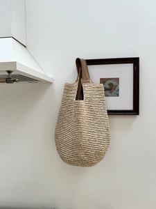 Close-up of woven straw shoulder bag with brown leather handle hanging on a white wall