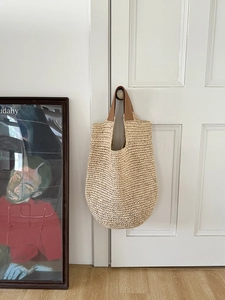 Detail of hand-woven straw shoulder bag with brown leather handles hanging on a white door