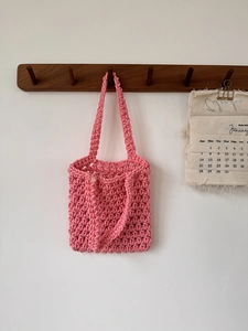 Pink hand-woven mesh tote bag hanging on wooden hook with rustic calendar background