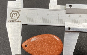 Macro shot measuring orange goldstone teardrop pendant width with a metal caliper