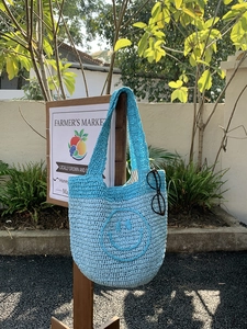 Detail of blue crochet tote bag with smiley face pattern hanging in a market scene