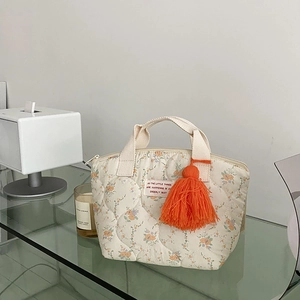 Détail du sac bento matelassé à fleurs avec pompon orange et étiquette sur étagère verre