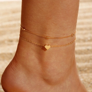 Detail of layered gold anklet with heart charm on sandy beach under sunlight