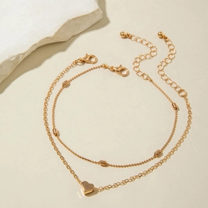 Close-up of golden layered anklet featuring heart pendant and metallic beads on a warm beige background