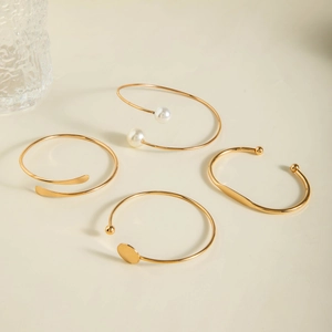 Detail of four gold-plated open bangles with pearls and geometric charms on cream surface