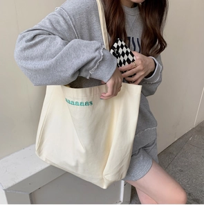 Close-up of beige canvas tote bag with green embroidered aaaaas lettering on the shoulder
