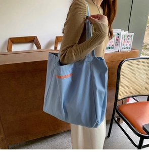 Detail of light blue canvas tote bag with orange maaaaaas embroidery on a female model