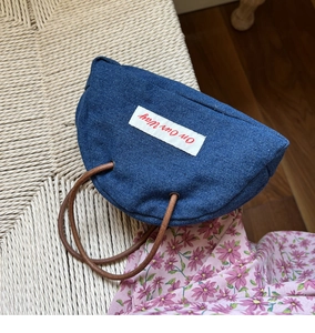 Detail of denim handbag with On Our Way text and brown cord handles in lifestyle setting