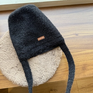 Black knit aviator hat with long earflaps and speckled texture on brown plush mat