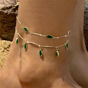 Detail of layered gold anklet with green marquise stones worn on an outdoor sandy beach