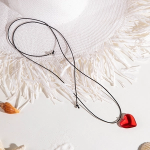 Detalle de collar con colgante de corazón rojo en arena de playa con conchas y sombrero