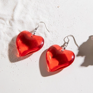 Macro view of transparent red heart pendant earrings with silver hooks on white sand