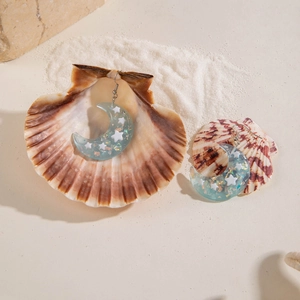 Close-up of resin crescent moon earrings with star sequins on a seashell and sand background