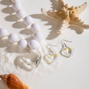 Détail d'un ensemble de bijoux style plage avec pendentifs cœur sur fond de sable blanc