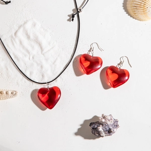 Detail of red translucent heart pendant necklace and earrings on white sand with shells