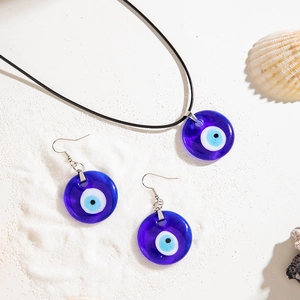 Detailed close-up of blue glass evil eye pendant and earrings on a white beach sand background