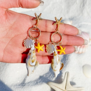 Golden starfish and conch drop earrings held over white sand for a beach vacation look