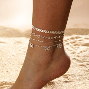 Detalle de tobilleras bohemias de plata con dijes de mariposa sobre la arena de playa