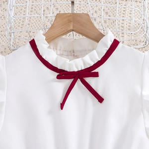 Detail of ruffle collar with red trim and bow on a white girls' dress hanging on a wooden rack