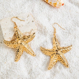 Detailed close-up of gold starfish earrings with textured surface on beach sand