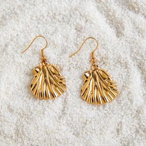 Détail de boucles d'oreilles coquillage dorées sur fond de sable blanc fin
