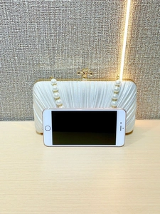 Size comparison of white pleated evening clutch with pearl handle and smartphone