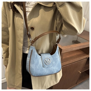 Detail of light blue denim half-moon bag with brown leather trim and silver M-letter buckle