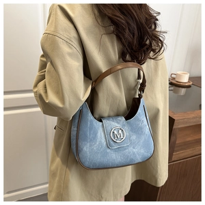 Détail du sac sous l'aisselle en denim bleu avec bordure en cuir marron et logo M métallique