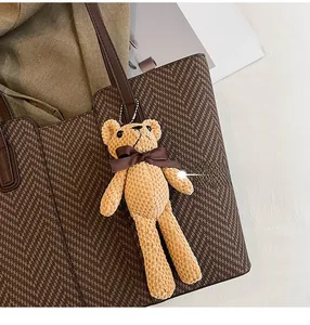 Close-up of a light brown plush bear charm with dark silk bow on a textured tote bag