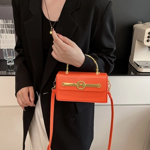 Orangefarbene Box-Tasche mit goldenem Griff und M-Logo-Verschluss auf schwarzem Blazer