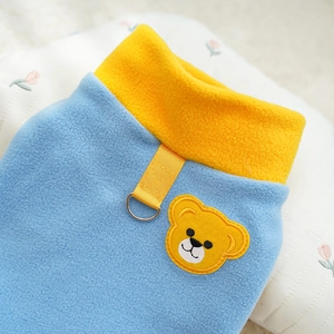 Detail of blue polar fleece pet coat featuring yellow bear embroidery and metal D-ring hook
