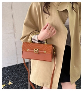 Close-up of a brown flap handbag with gold metal M logo and handle in an outdoor setting