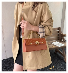 Detail of a brown leather box bag featuring a gold M logo and elegant metallic top handle