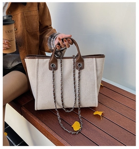 Detail of canvas tote bag with brown leather trim and metal chain strap on a wooden bench