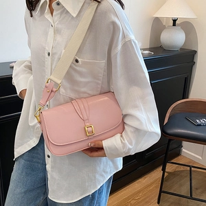 Pink crossbody bag with wide strap and gold buckle shown in a minimalist room